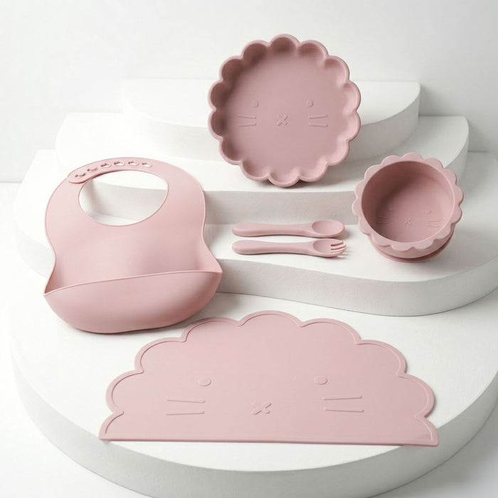 Beikost-Set aus Silikon in Rosa mit Löwengesicht: Lätzchen, Teller, Schale, Tischset, Löffel und Gabel, auf weißen Stufen