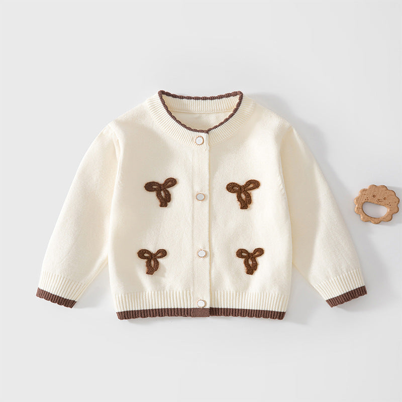 Cremefarbene Strickjacke mit braunen Schleifen neben einem Holzspielzeug